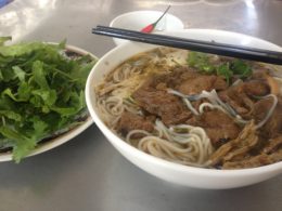 Photo contains a bowl of noodles with veg beef, mushrooms, sprouts, and other vegetables. On a separate plate, there are fresh herbs and leafy greens.