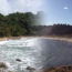 Image contains two beaches blending into one another in the middle of the picture. The beach on the left has sand that arches upward to the right, and there are palm trees lining the shore. The beach on the right has sand that arches upward to the right where it meets the edge of the other beach. Along the edge of the beach, there are sand dunes and evergreen trees with a few people walking on the shore.