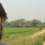 Image contains a photo of a man on the left-hand side wearing a face mask over his mouth and nose. To the right of the man, a hazy sky is visible above some trees in the horizon, a green rice field, and a narrow road.
