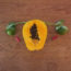 Image contains a medium-dark brown wooden background. On the wood, there is a variety of fruit and vegetables. In the centre, there is a papaya and coming from the left and right of the fruit are two small stalks of a leafy green vegetable. Nestled under the leaves at the end of each stalk are lemons, and just below each lemon on the right and left there is a small hot pepper. The whole image is meant to represent the female reproductive system.