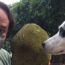 Image contains a large, leafy green tree in the background. In the foreground, there is a man on the left wearing a face mask. The man is holding a large spiny light green fruit to his right, and there is a dog with black patches around her eye that is looking directly at the man.