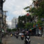 Image is a photo of a Saigon street with numerous scooters coming towards the camera. Shops are visible on both sides of the street, and a small neon sign in the top left-hand corner says "Quán Chay Giác Tha".