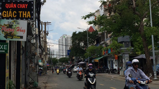 Image is a photo of a Saigon street with numerous scooters coming towards the camera. Shops are visible on both sides of the street, and a small neon sign in the top left-hand corner says "Quán Chay Giác Tha".